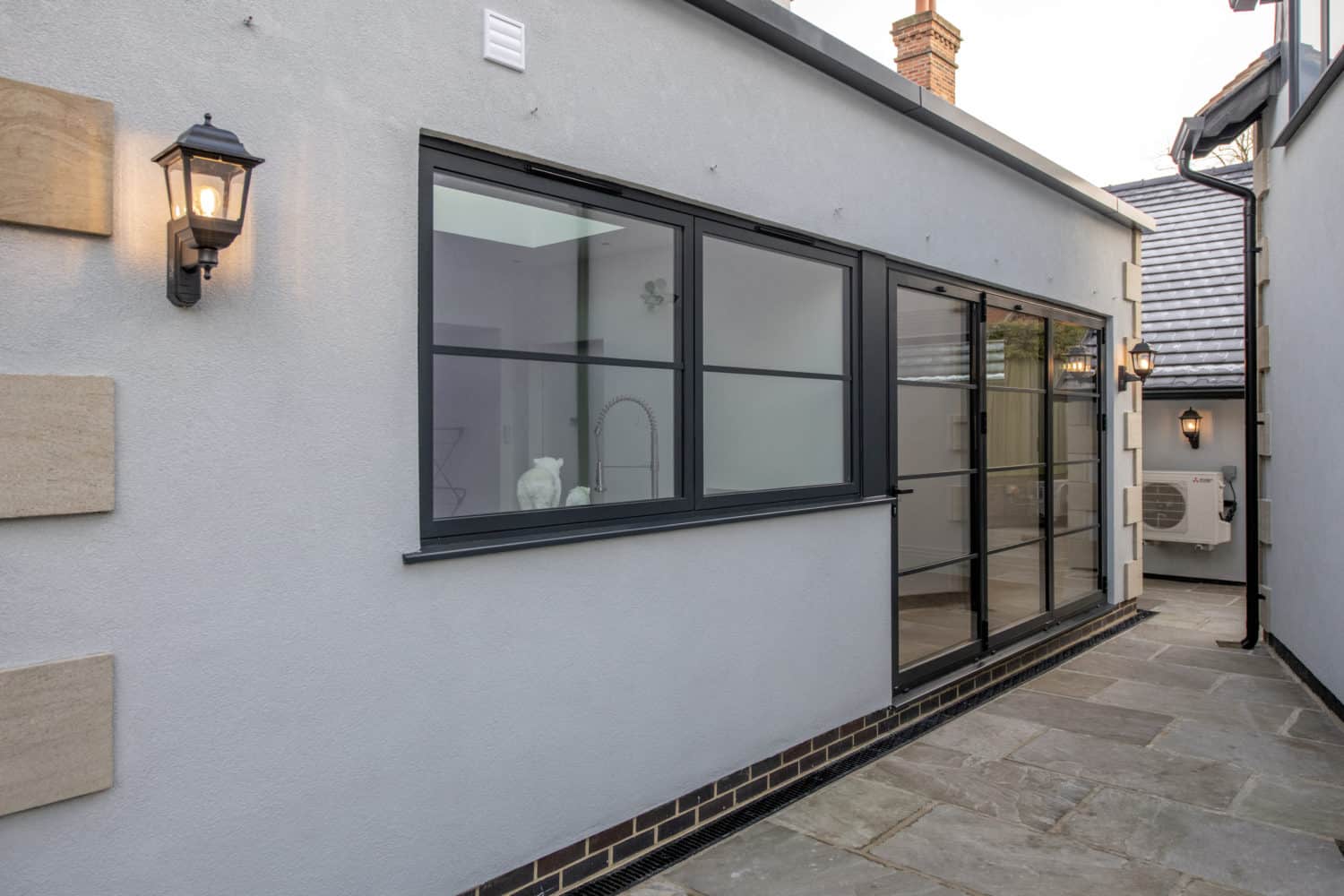 Modern exterior of a house with light gray walls, black-framed windows, bifold doors, outdoor wall lights, and a stone-tiled patio. A kitchen sink and faucet are visible through the window.
