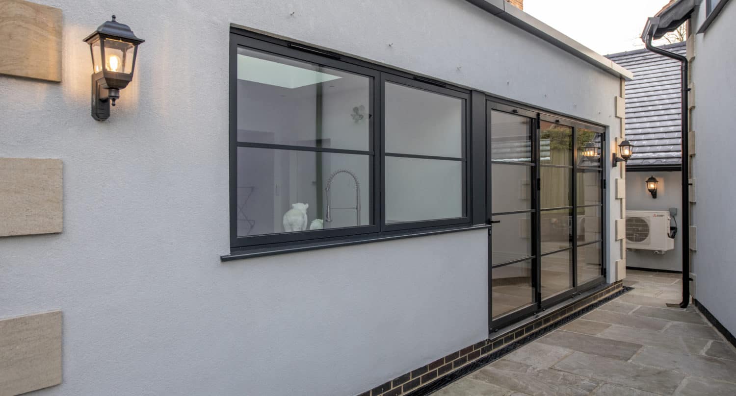 Modern exterior of a house with light gray walls, black-framed windows, bifold doors, outdoor wall lights, and a stone-tiled patio. A kitchen sink and faucet are visible through the window.