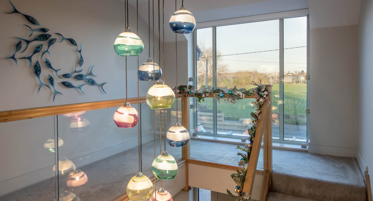 A modern stairway with glass railings and leafy garland sits beneath colorful pendant lights. Bespoke glass solutions are showcased in the fish wall art and large windows, which overlook a green field outside.