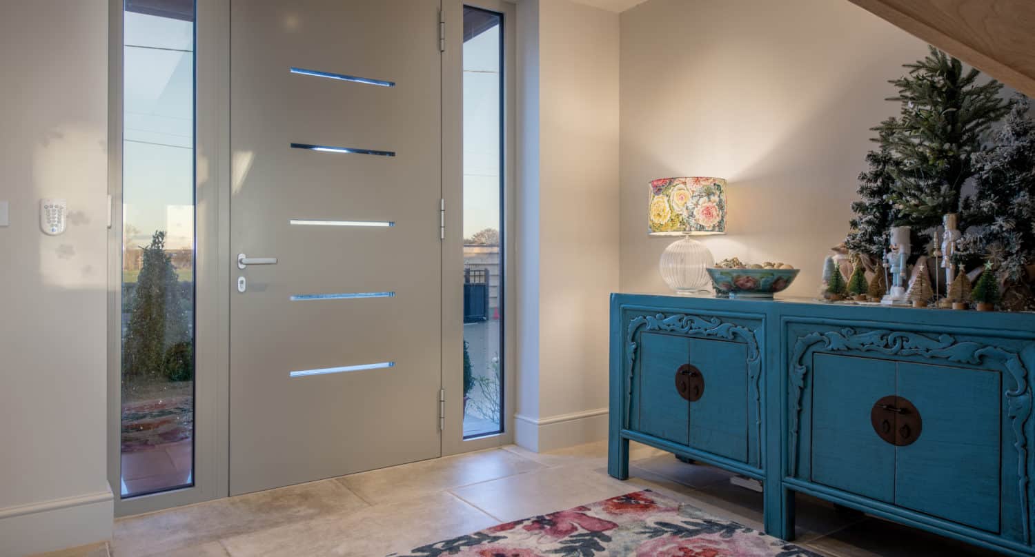 A modern entryway with a light gray front door featuring horizontal glass panels, a floral rug, a blue sideboard with decorative items, and a lamp with a colorful shade. Light streams in through the side window.