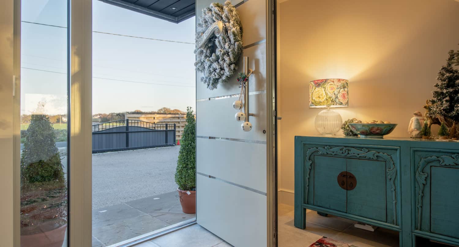 A modern front door decorated with a frosted wreath is open, revealing an entryway with a blue cabinet, colorful lamp, and holiday decor. Outside, potted plants frame the front doors and offer a view of the driveway and neighborhood.