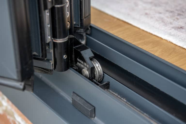 Close-up view of a sliding door roller mechanism fitted into a metal track, perfect for smooth-operating sliding doors, with part of a wooden floor and light-colored rug visible in the background.
