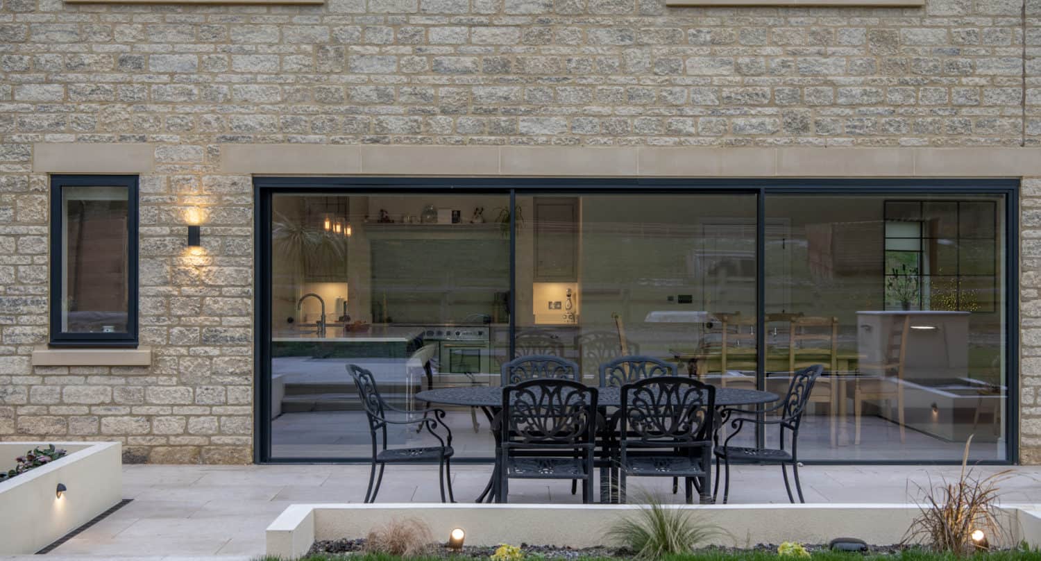 A modern patio with black metal table and chairs sits outside a house with large sliding doors, revealing a bright kitchen and dining area inside. The exterior wall is made of light stone bricks.