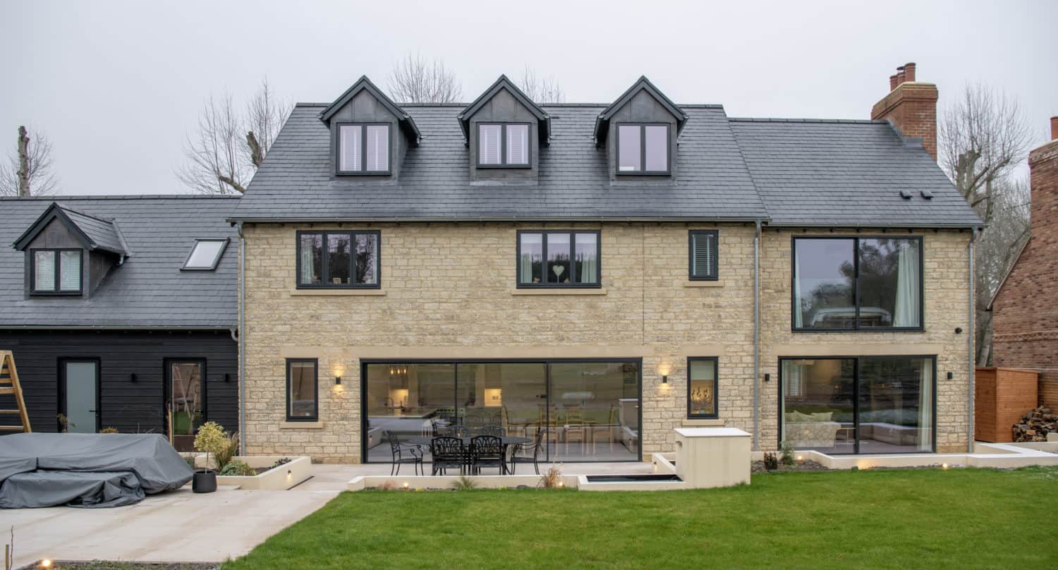 Modern stone house with black-framed windows, bespoke glass solutions including a large sliding door opening to a patio with outdoor furniture, and a green lawn in the foreground. Multiple dormers add to its contemporary charm.