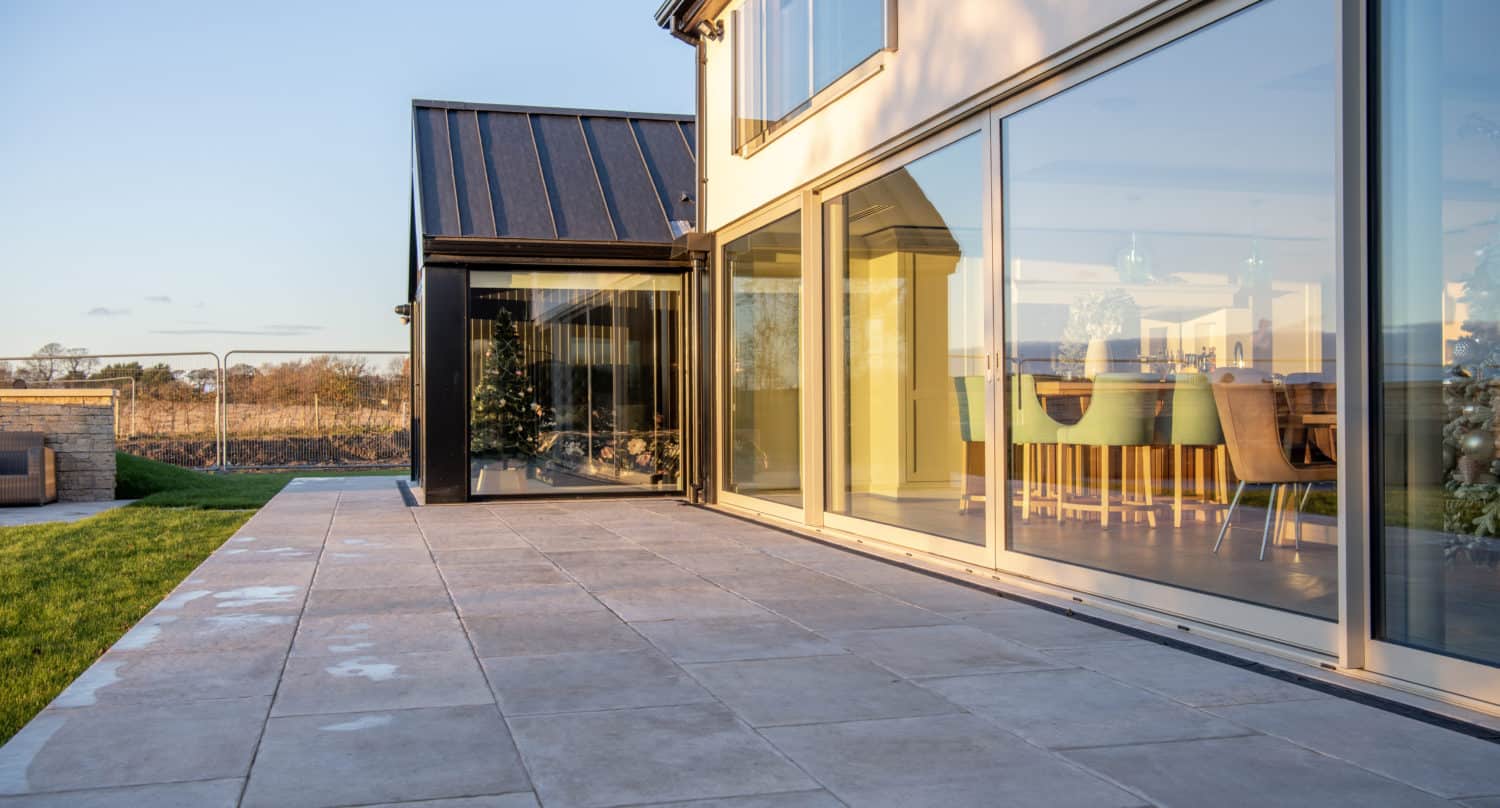 A modern house with large bifold doors opens onto a spacious tiled patio. Inside, a dining table and chairs are visible. The patio overlooks a grassy lawn and open landscape under a clear sky.