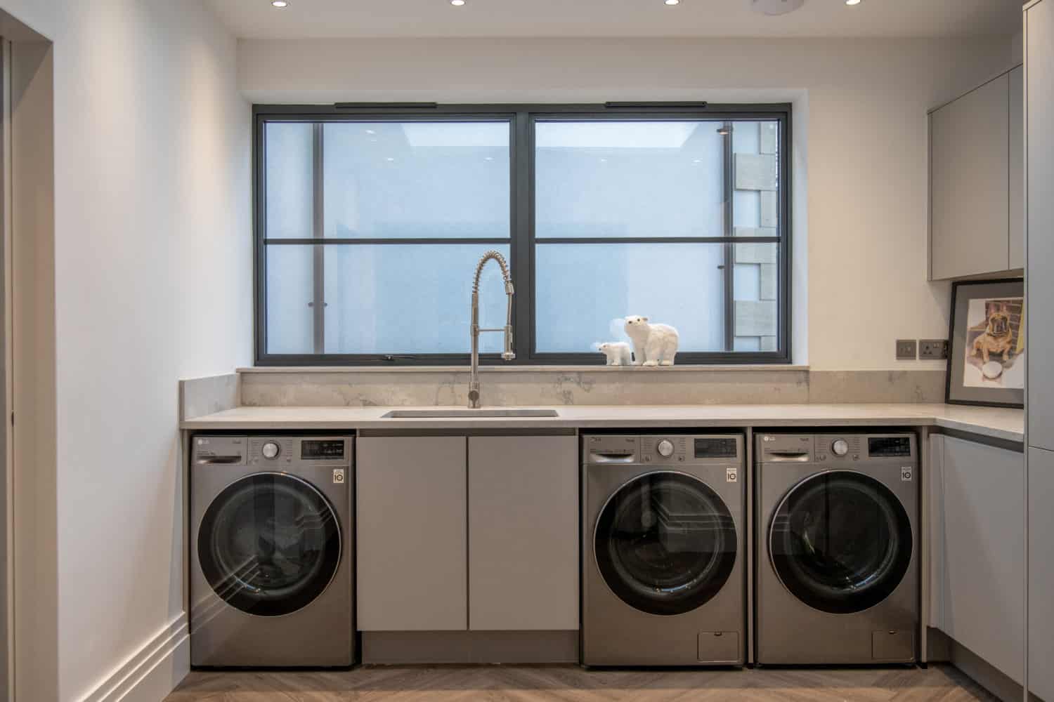 Modern laundry room with gray cabinets, a large sink with a tall faucet, two washing machines, two dryers, and a big window. Bifold doors open to the outdoors. A white polar bear figurine and a framed photo are displayed on the countertop.