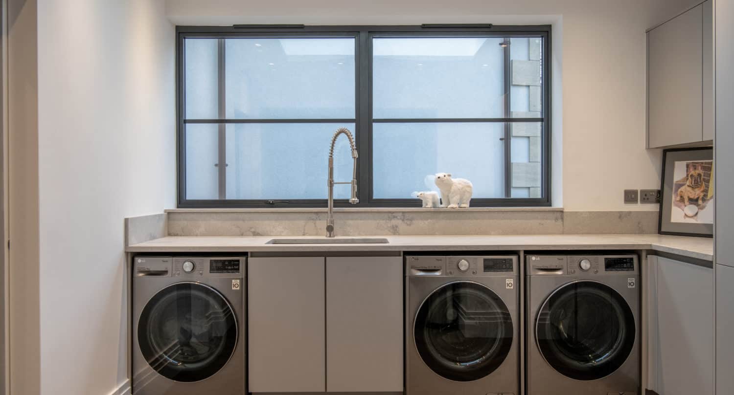Modern laundry room with gray cabinets, a large sink with a tall faucet, two washing machines, two dryers, and a big window. Bifold doors open to the outdoors. A white polar bear figurine and a framed photo are displayed on the countertop.