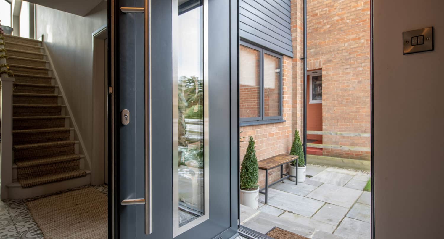 A modern front door with a large glass panel is open, revealing a stone patio with two potted plants and a wooden bench outside. Inside, sliding doors lead to a tiled floor and a staircase with carpeted steps.