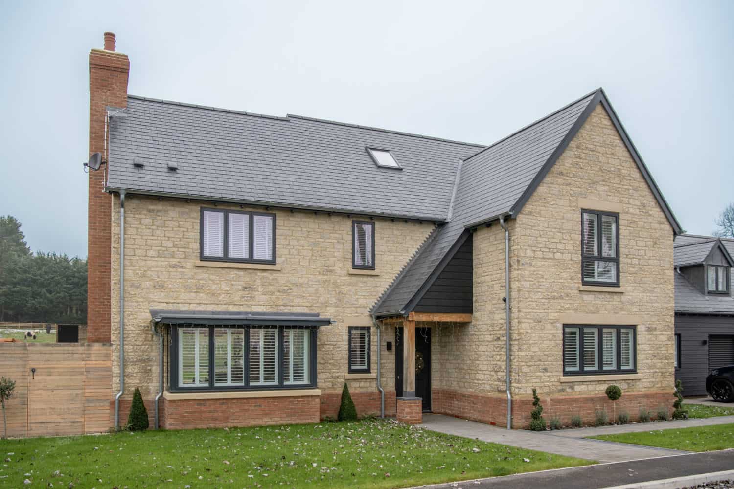 A modern two-story house with beige brick walls, gray roof, and large windows with white shutters. Bifold doors open onto the front yard, which features a green lawn and a small pathway leading to a covered front entrance.