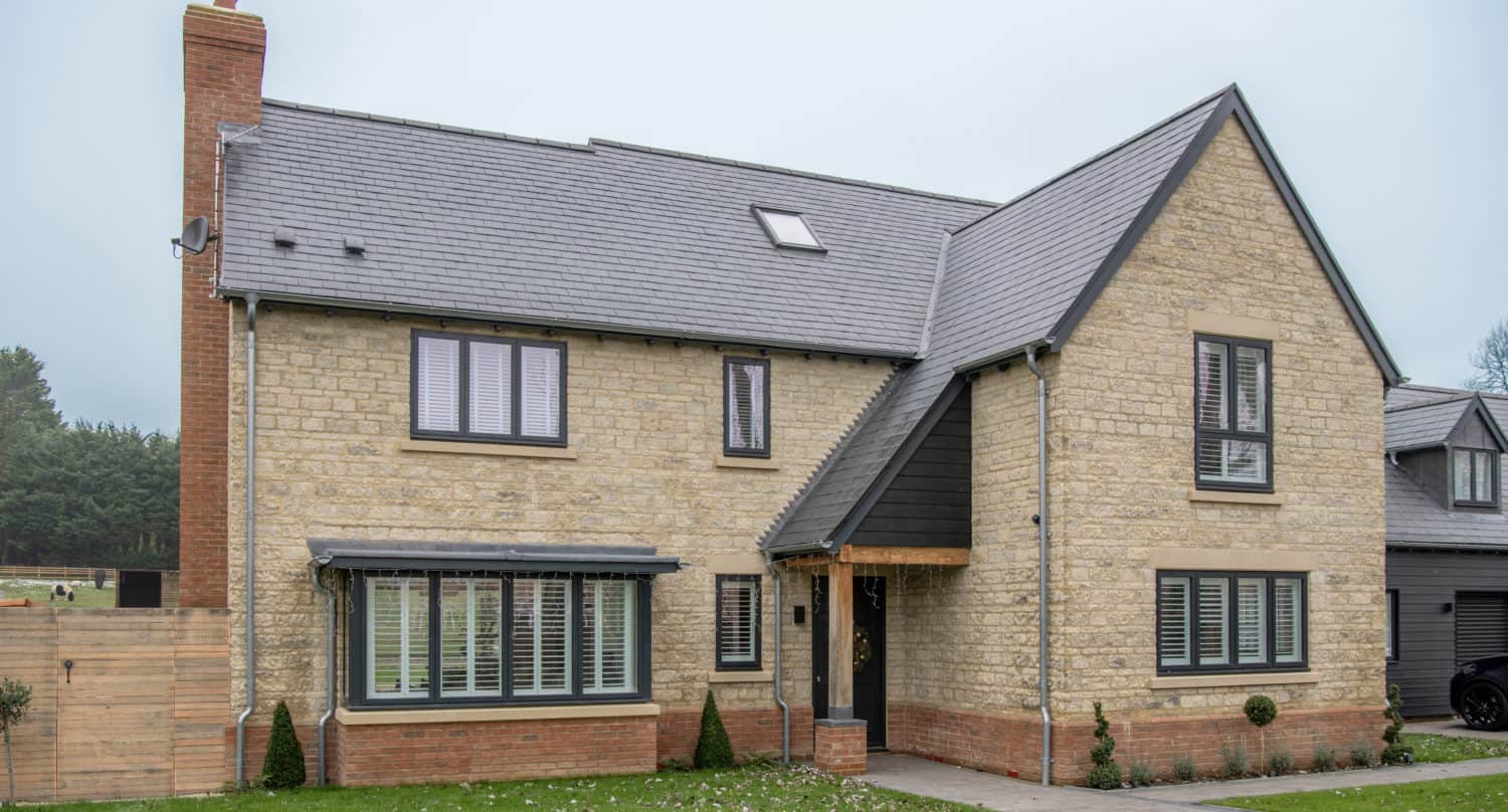 A modern two-story house with beige brick walls, gray roof, and large windows with white shutters. Bifold doors open onto the front yard, which features a green lawn and a small pathway leading to a covered front entrance.
