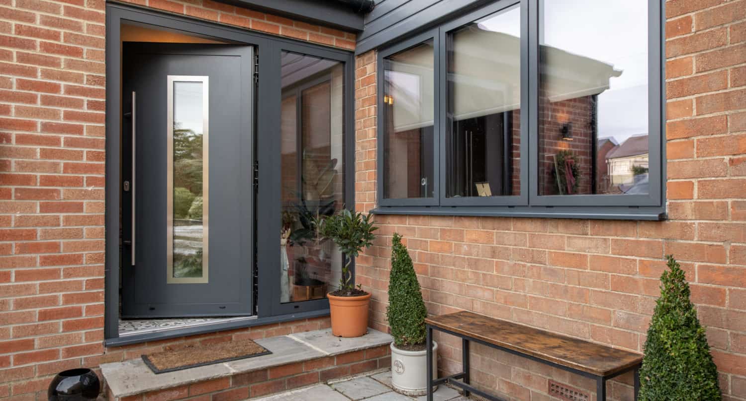 Modern brick house entrance with a dark gray front door, large window, welcome mat, potted plants, and a wooden bench on a stone patio beneath stylish glass roofing.