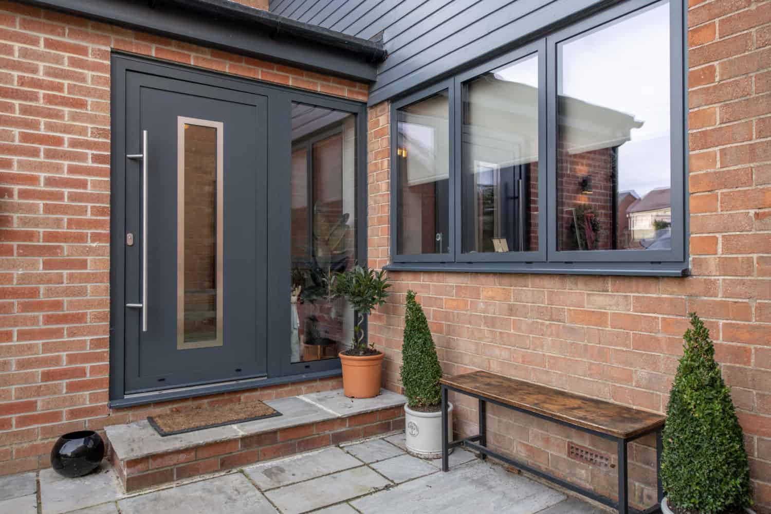 A modern brick house entrance with a dark gray front door, large window, potted plants, a doormat, a wooden bench on a stone patio, and sleek sliding doors that open to the outside.