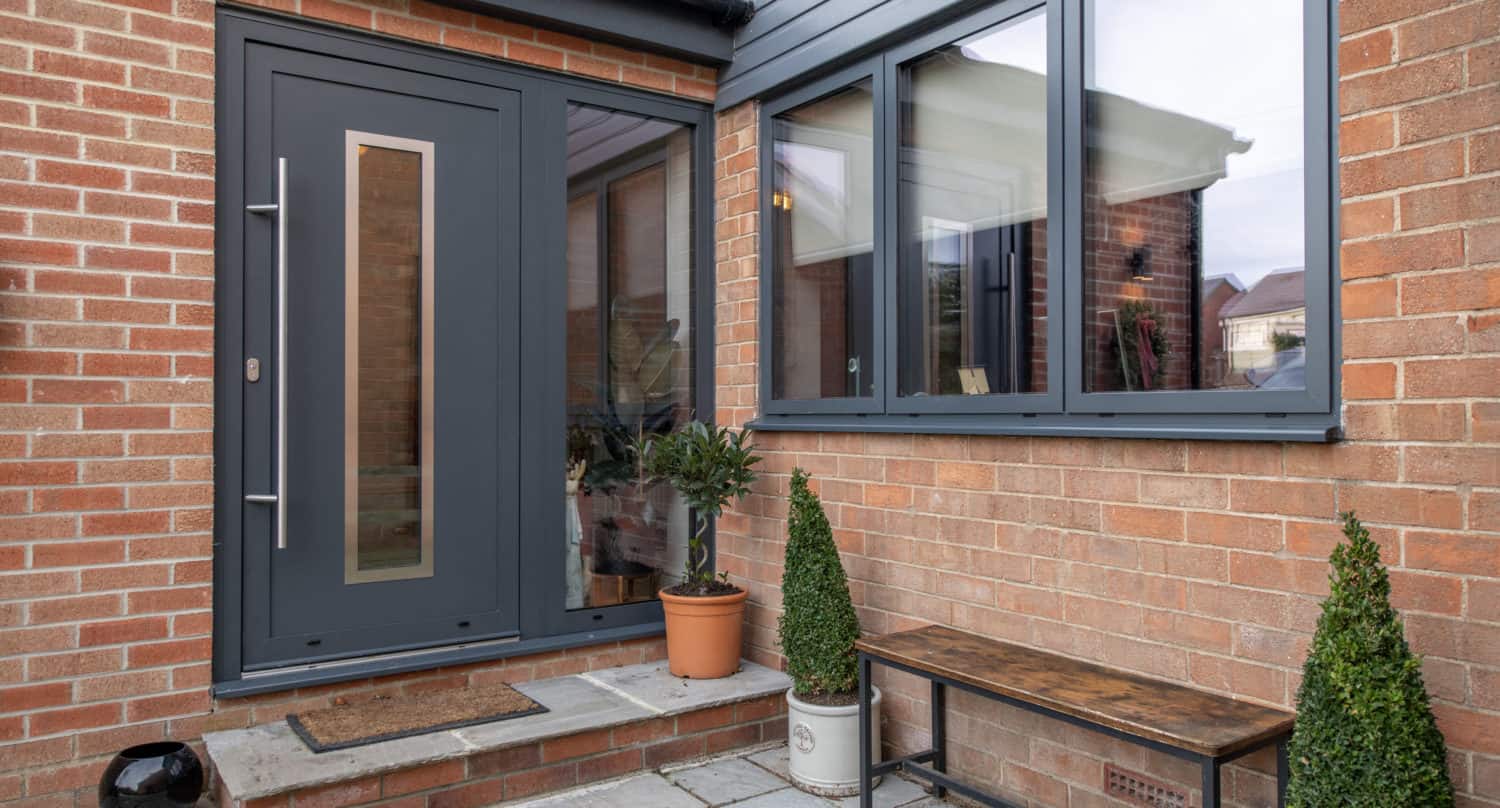 A modern brick house entrance with a dark gray front door, large window, potted plants, a doormat, a wooden bench on a stone patio, and sleek sliding doors that open to the outside.