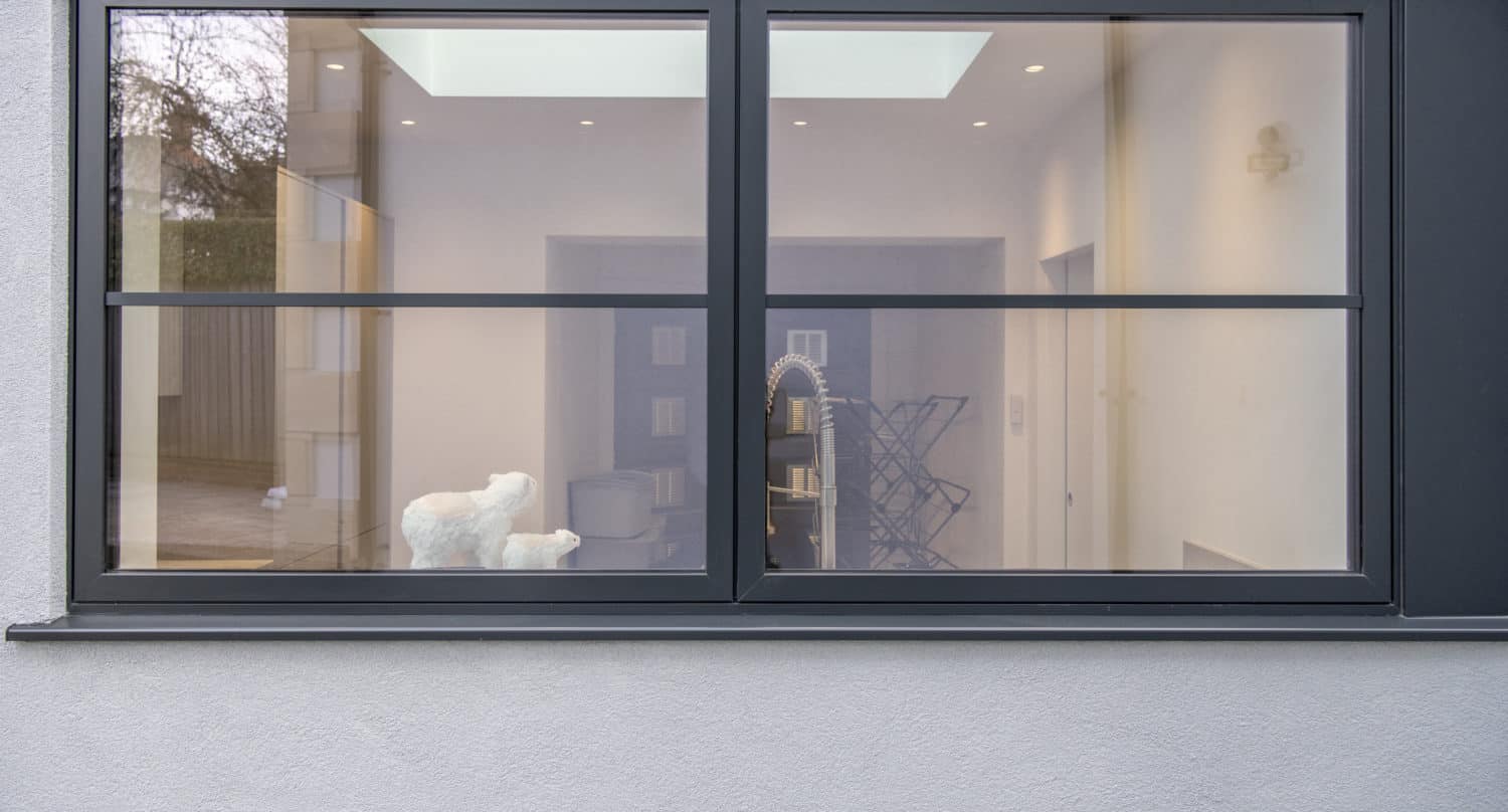 Large double-pane window on a gray wall, showing a modern interior with white walls, a light fixture, skylight, and a sculpture of a white animal on a table inside. Reflections appear on the glass—perfect for showcasing bespoke glass solutions.