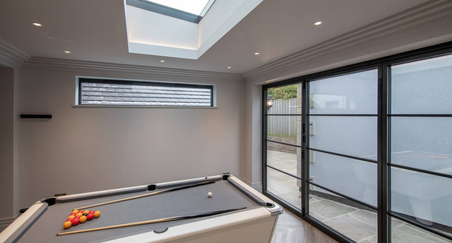 A modern room with a white pool table set up for a game, featuring glass roofing above and large bifold doors on the right side letting in natural light.
