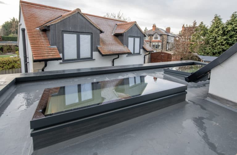 A flat roof with large, rectangular rooflights and standing water, overlooking a house with pitched red-tile roofs, grey-framed windows, and sleek sliding doors on an overcast day.