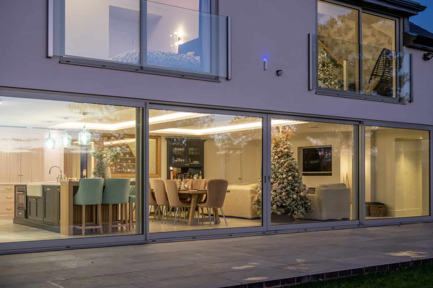 Modern home with large glass windows and sleek sliding doors reveals a kitchen, dining area, and living room adorned with a Christmas tree. Warm indoor lighting creates a cozy contrast against the evening light outside.