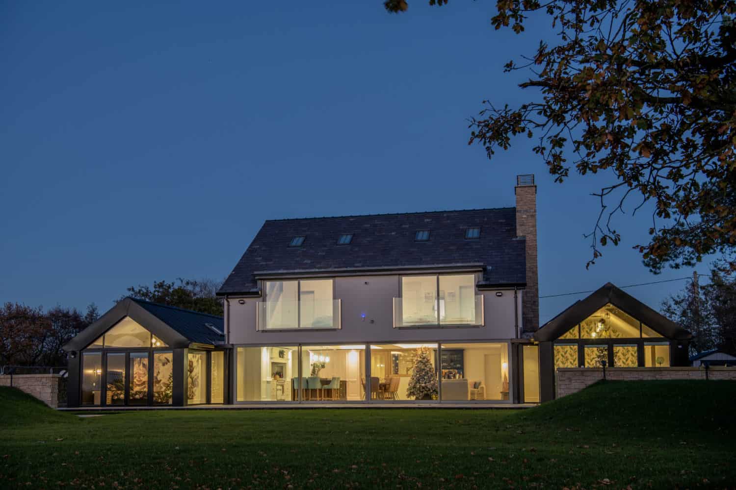 A modern two-story house with large glass windows and bifold doors, warmly lit from inside, sits on a spacious lawn at dusk with trees in the background.