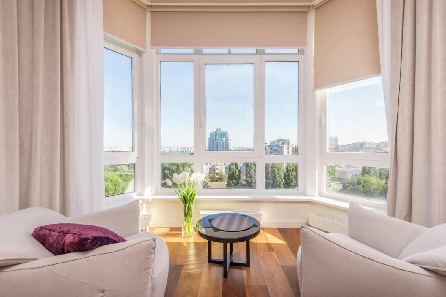 Bright room with two white armchairs, a small round table, wood flooring, and bespoke glass solutions in large corner windows reveal a stunning cityscape view under a clear sky. Cream curtains and white tulips complete the inviting scene.