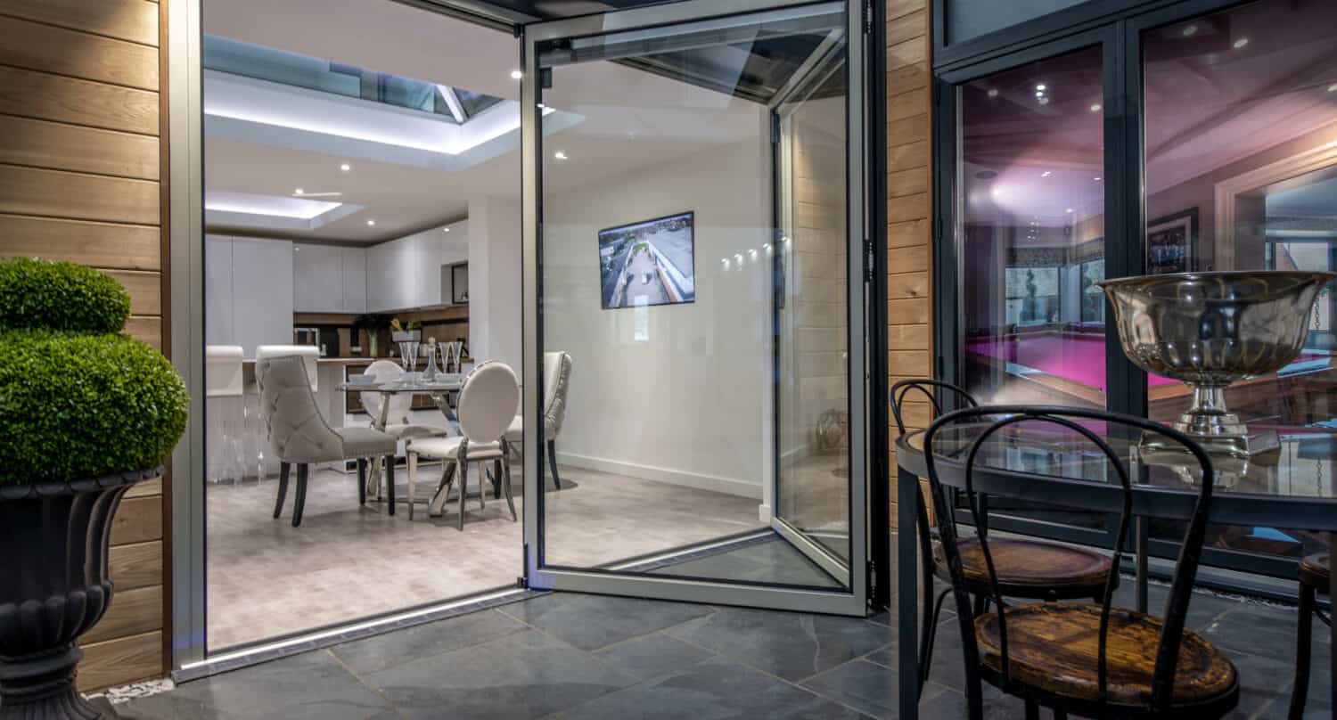 Modern dining area with bifold doors opening to a patio. Indoor space features a dining table, white chairs, and a TV on the wall. Outdoor area has a metal table, chairs, and potted plant on a stone floor.