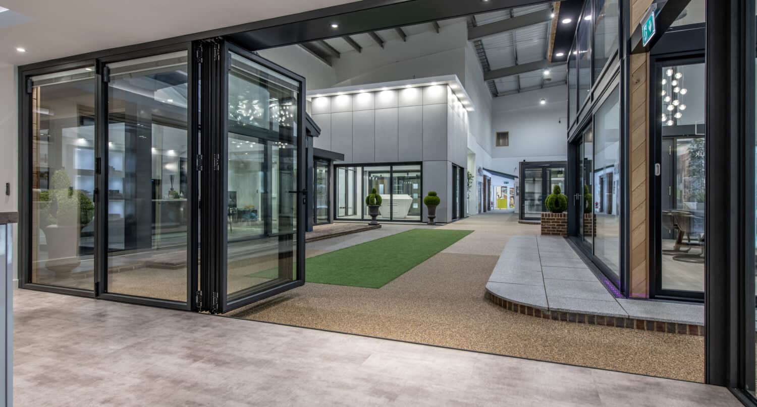 Modern indoor showroom with large bifold doors, green artificial grass, potted plants, and various display rooms under industrial ceiling lights. The space appears open, sleek, and well-lit with bespoke glass solutions throughout.