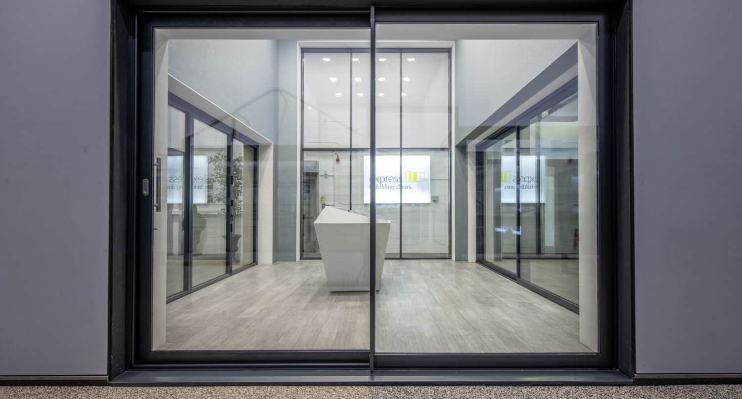 A modern glass sliding door opens to a sleek, minimalist office lobby with light wood floors, white walls, and a white angular reception desk in the center. Bifold doors complement the design, while digital signage is visible at the back.