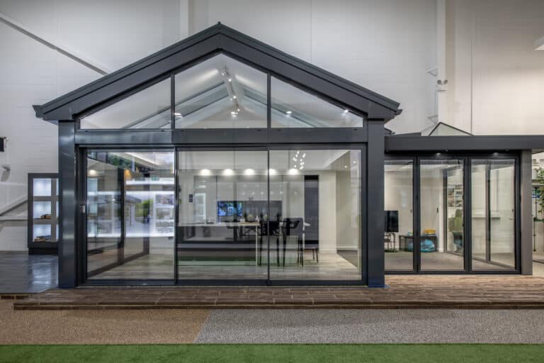 Modern glass and metal structure with a gabled roof, featuring large bifold doors and windows, situated indoors with views of a showroom interior and displays behind.