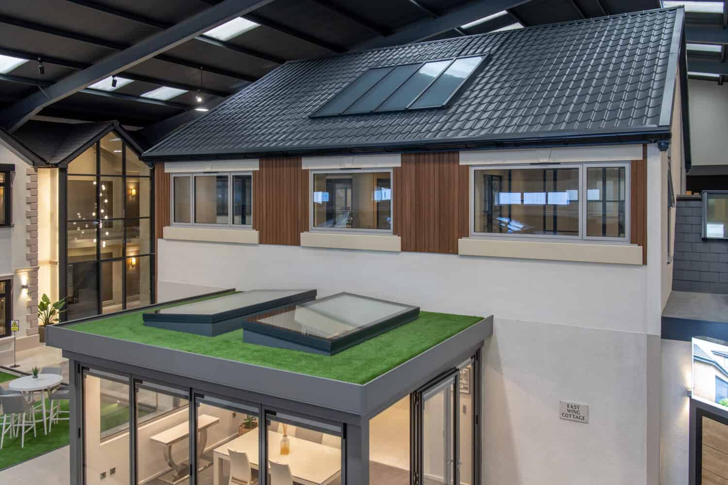 A modern house display inside a showroom features large windows, a glass extension with skylights, bifold doors opening to a green rooftop terrace, and a sloped roof with solar panels under a high ceiling.