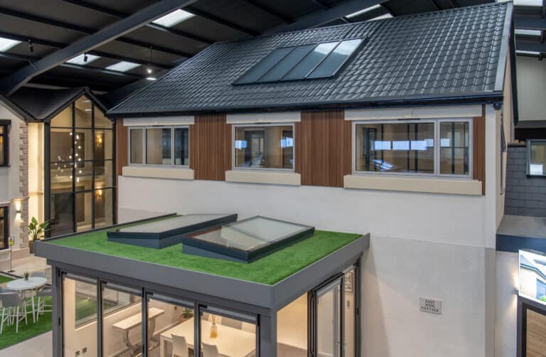 A modern house display inside a showroom features large windows, a glass extension with skylights, bifold doors opening to a green rooftop terrace, and a sloped roof with solar panels under a high ceiling.