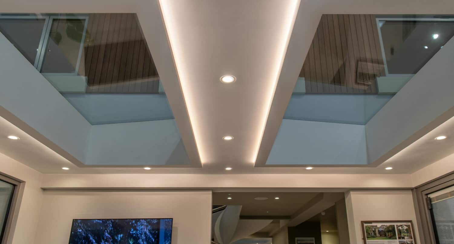 Modern interior ceiling with recessed lighting, two large rectangular skylights, and soft indirect LED lights outlining the skylights. Bespoke glass solutions complement the framed art and TV visible on the walls below.