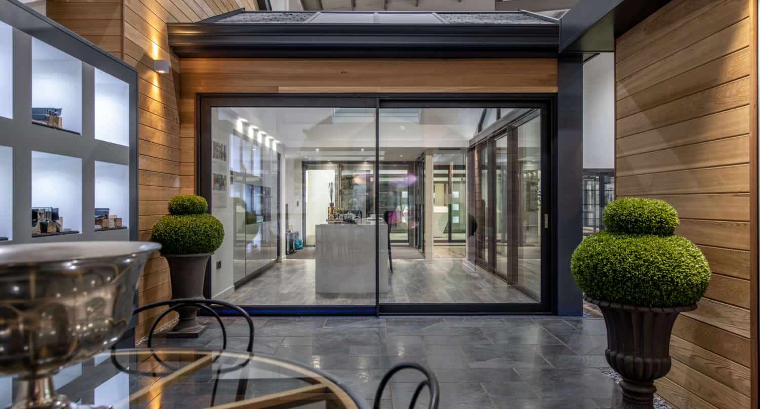 Modern indoor space featuring a glass table with chairs, two round topiary plants, and bespoke glass solutions like large sliding doors leading to a stylish interior with wooden and metal elements and display shelves.