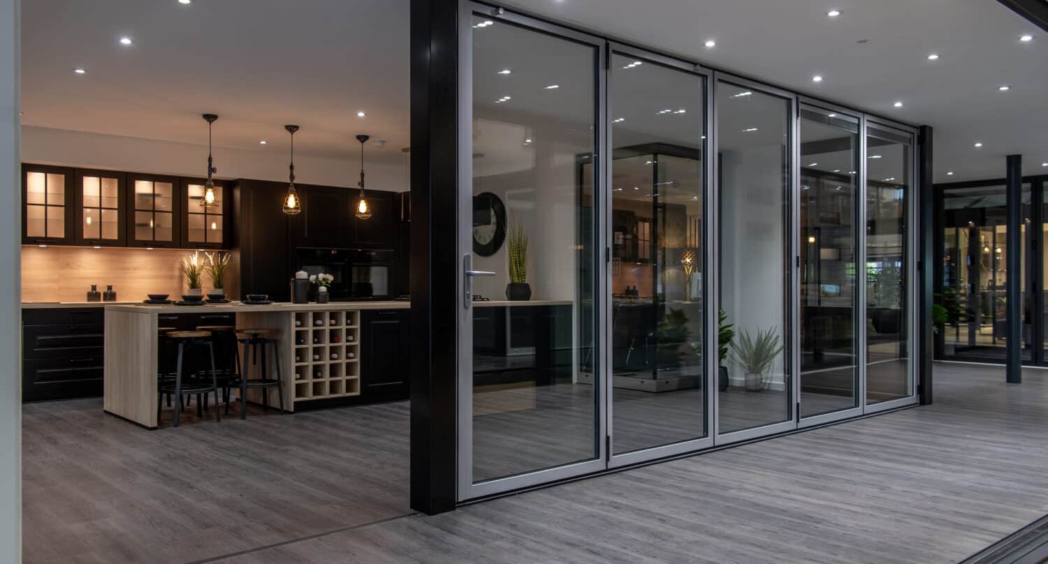Modern open-plan kitchen with dark cabinetry, pendant lighting, an island with bar stools, and floor-to-ceiling bifold doors opening to a spacious area with wood-style flooring.