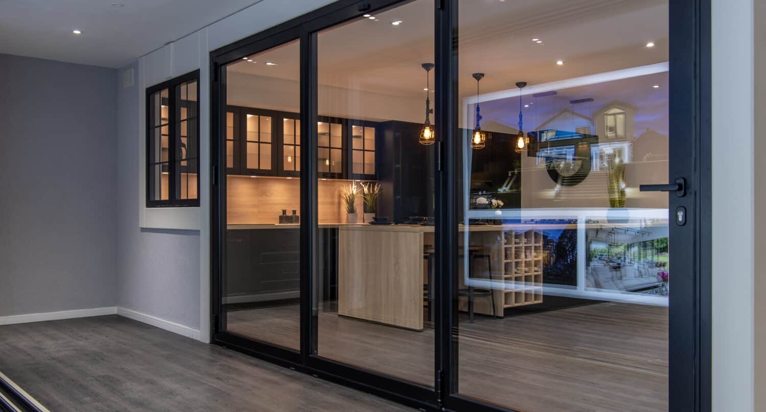 Modern kitchen viewed through large glass bifold doors; features black cabinets, a wooden island, pendant lights, and a wine rack, with reflections of an outdoor scene visible in the glass.