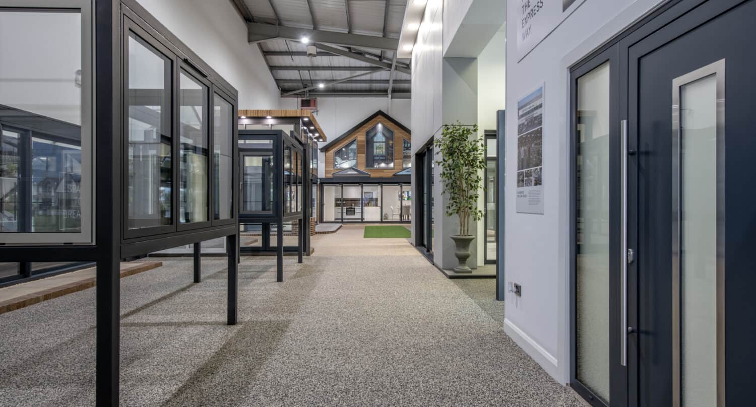 A modern showroom interior displaying large window and door frames, including sleek bifold doors, with a model house visible in the background and a tall indoor plant on the right side of the walkway.