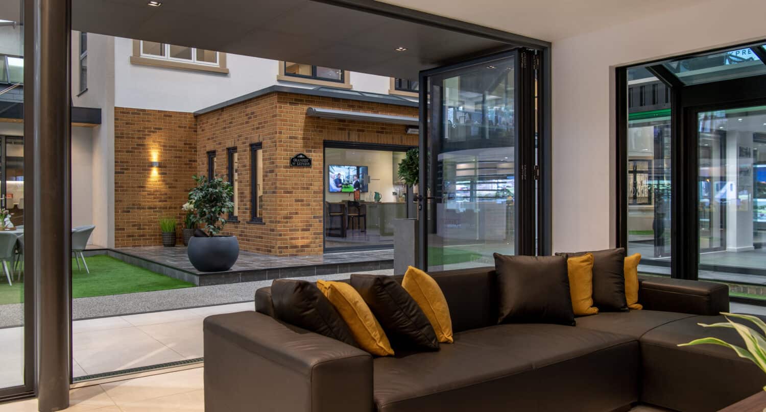 Modern living room with a dark sectional sofa and gold accent pillows, large glass doors open to a patio with artificial grass, potted plants, and a brick building in the background enhanced by bespoke glass solutions.