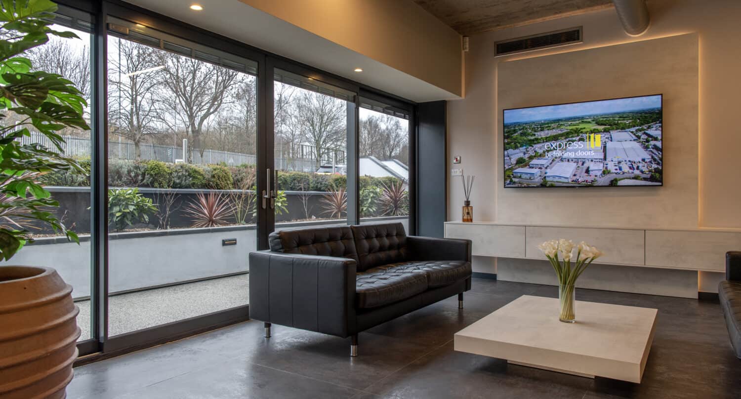 Modern lobby with black leather sofas, a light square coffee table with white flowers, large windows and bespoke glass solutions offering views of the outdoor area with plants, and a wall-mounted TV displaying an aerial image.
