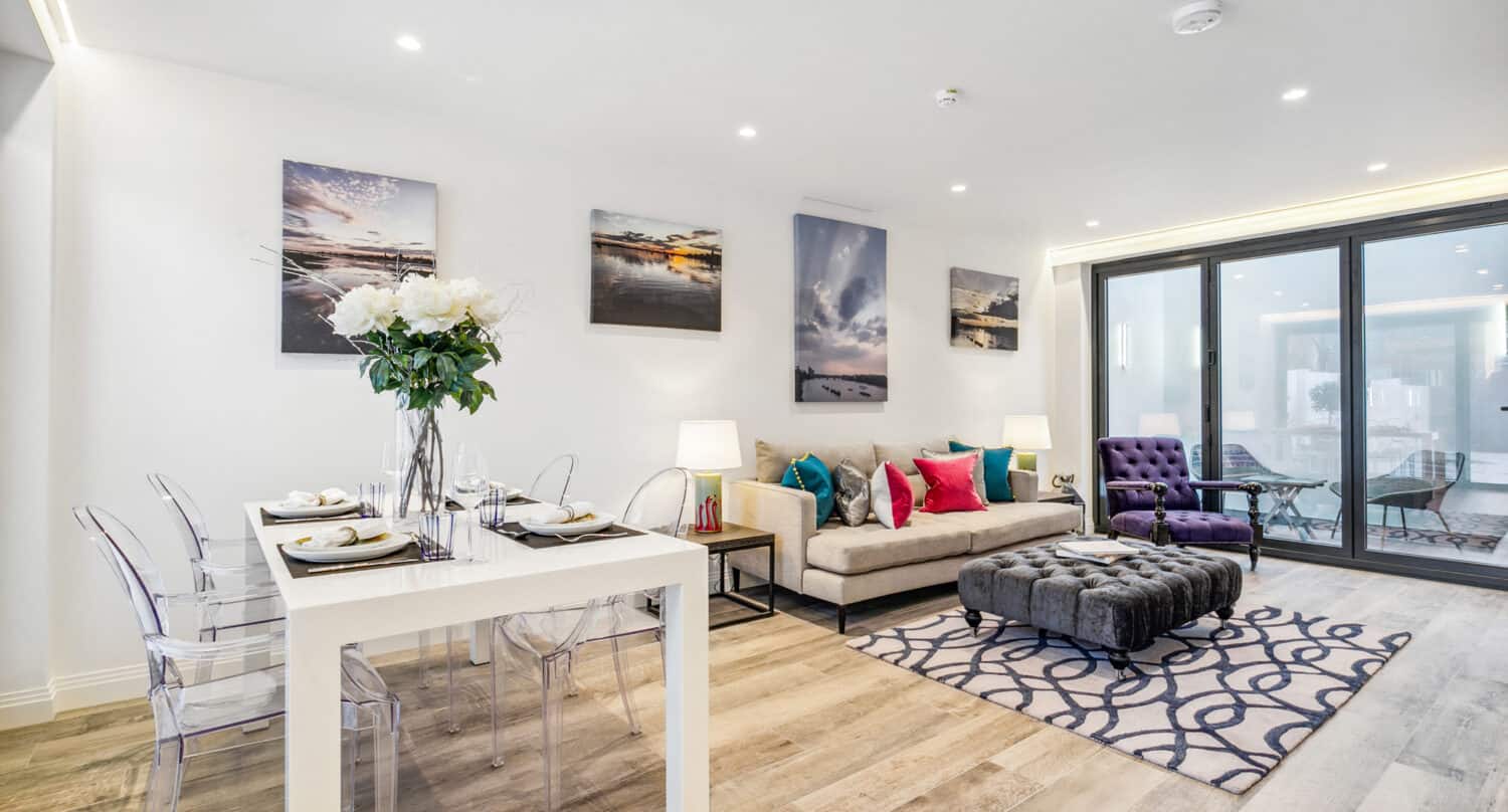 Modern open-plan living room with a beige sofa, colorful cushions, a purple armchair, clear dining chairs, a white dining table, wall art, rug, and large bifold doors leading to a bright balcony.
