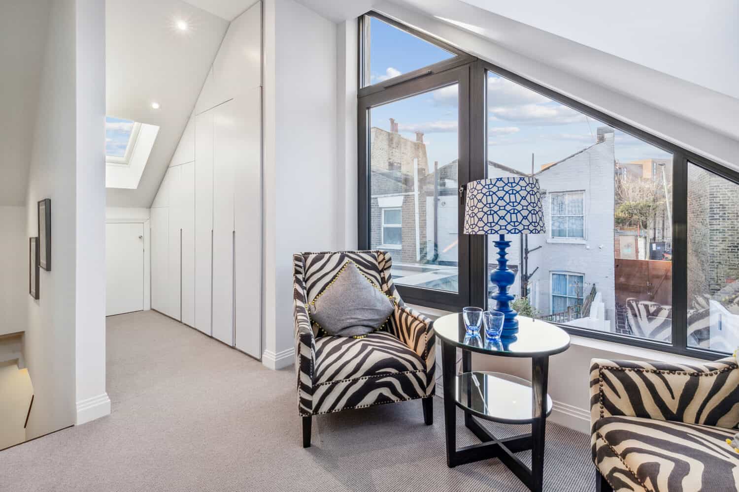 Modern sitting area with two zebra-print armchairs, a round glass table with a blue lamp, and large windows overlooking neighboring buildings; white walls and bespoke glass solutions like a skylight provide ample natural light.