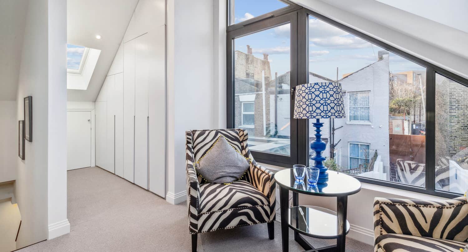 Modern sitting area with two zebra-print armchairs, a round glass table with a blue lamp, and large windows overlooking neighboring buildings; white walls and bespoke glass solutions like a skylight provide ample natural light.