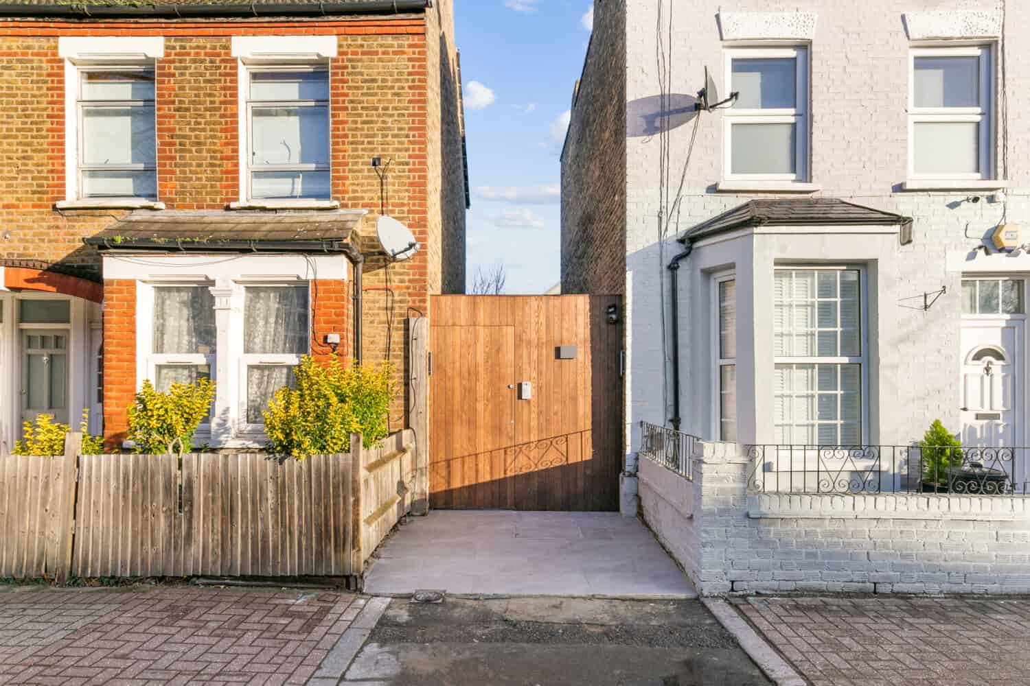A wooden gate stands between two attached houses—one with red brick and a wooden fence, the other painted white with a metal railing—filling a narrow gap that could be enhanced with bifold doors leading to a hidden area behind the houses.