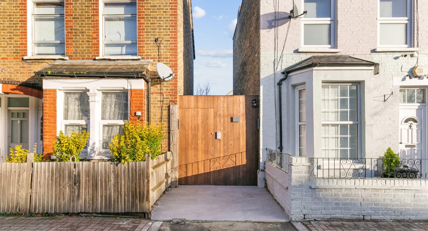 A wooden gate stands between two attached houses—one with red brick and a wooden fence, the other painted white with a metal railing—filling a narrow gap that could be enhanced with bifold doors leading to a hidden area behind the houses.