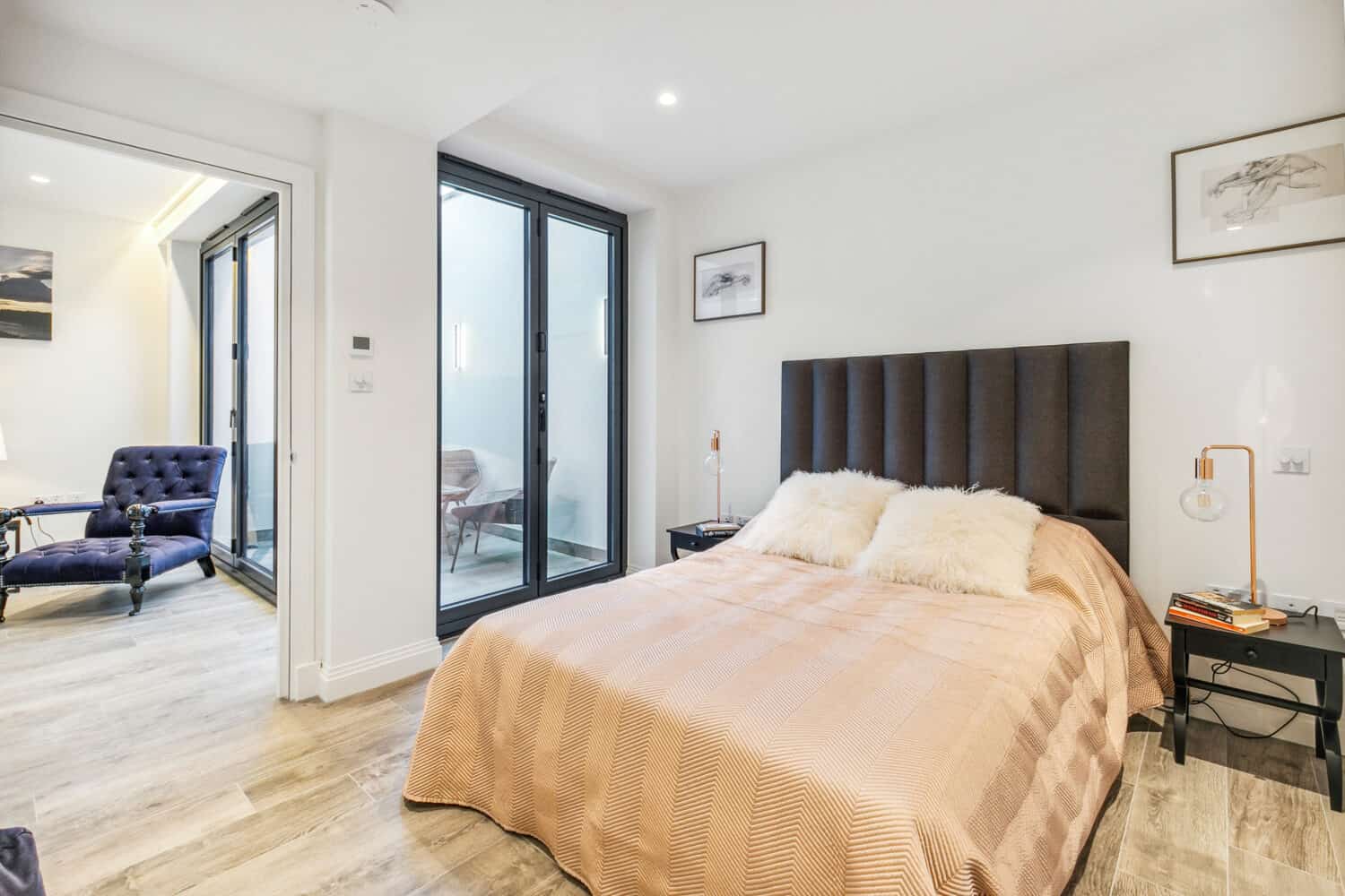 Modern bedroom with a double bed featuring a tan blanket and white fur pillows, two bedside tables, and sliding doors leading to a small sitting area with a blue chair. Walls are white with minimal artwork.