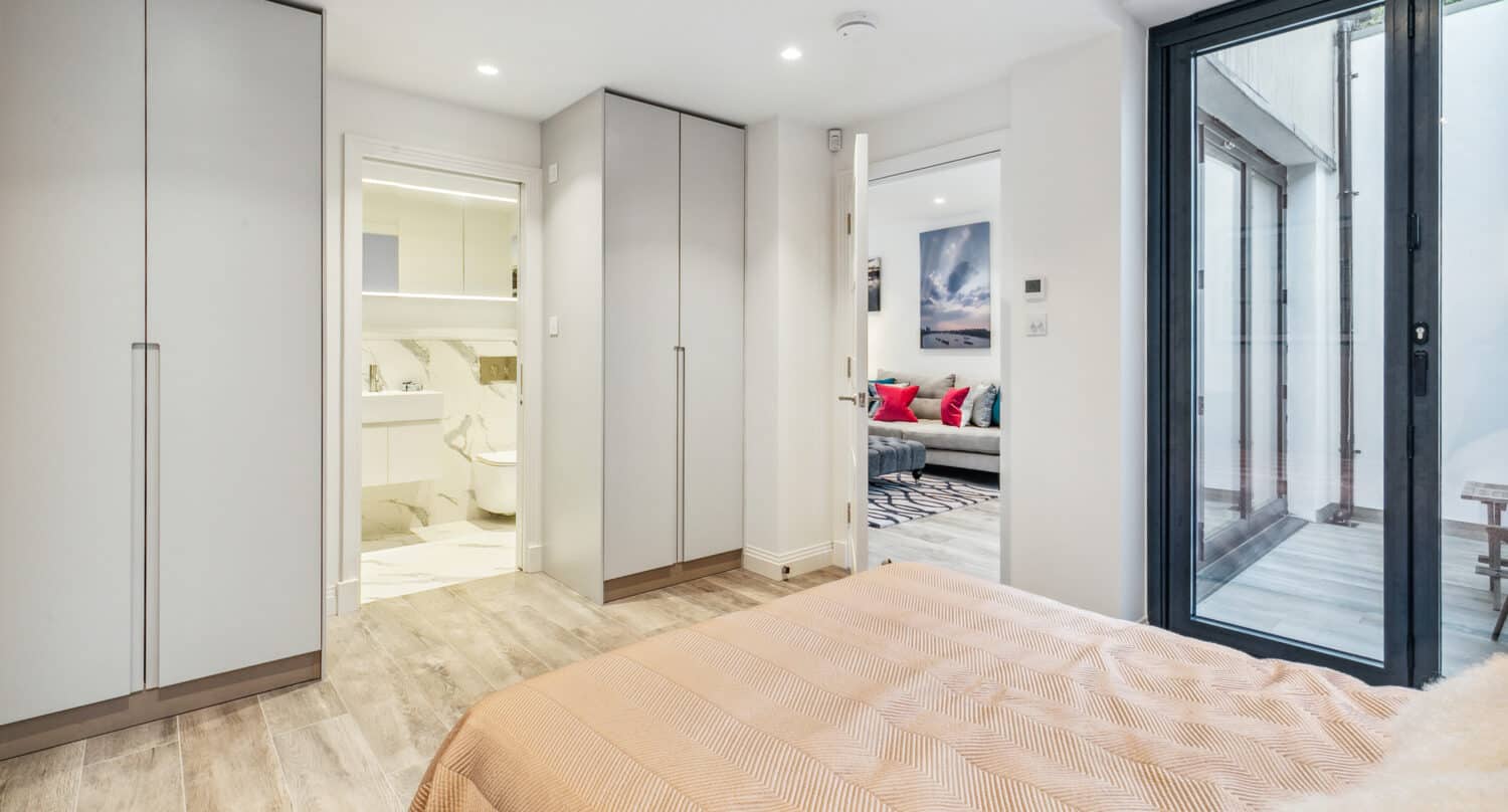 A modern bedroom with light wood flooring, a beige bedspread, built-in wardrobes, an en-suite bathroom, and bifold doors leading to an outdoor area. A living room with a sofa and colorful pillows is visible through an open door.
