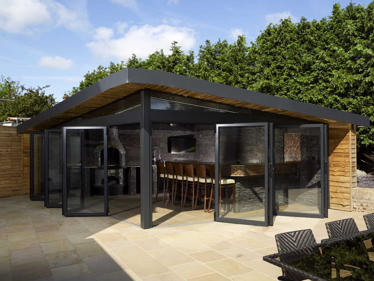 Modern outdoor garden room with sleek bifold doors open, bar-style seating, and a long wooden counter, set against tall green hedges and a clear sky. Patio area with seating is visible in the foreground.