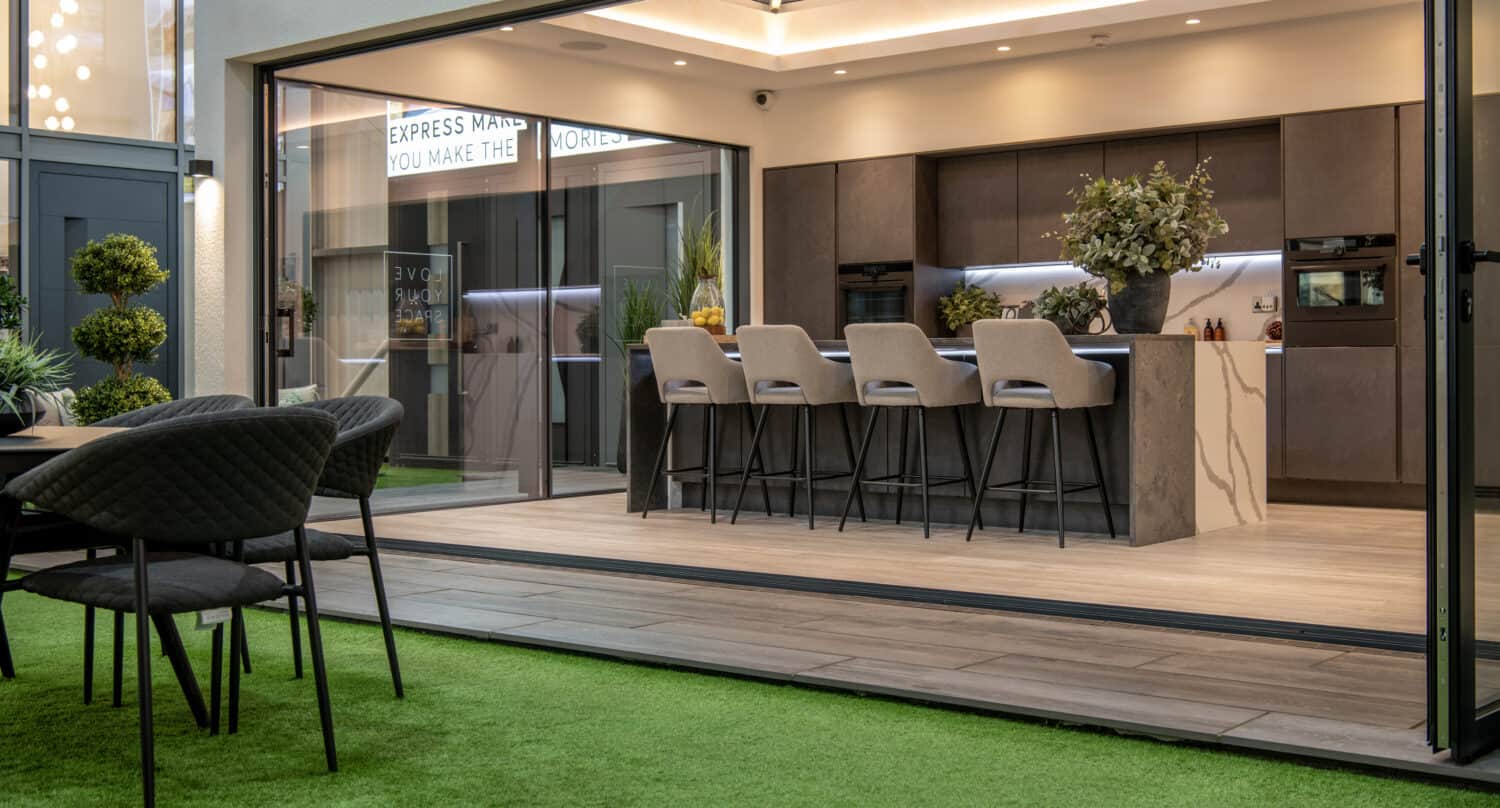 Modern open-plan kitchen with a large island and bar stools, seen through bespoke glass solutions like sliding doors from an outdoor patio area with artificial grass and a round dining table. Indoor and outdoor spaces blend seamlessly.