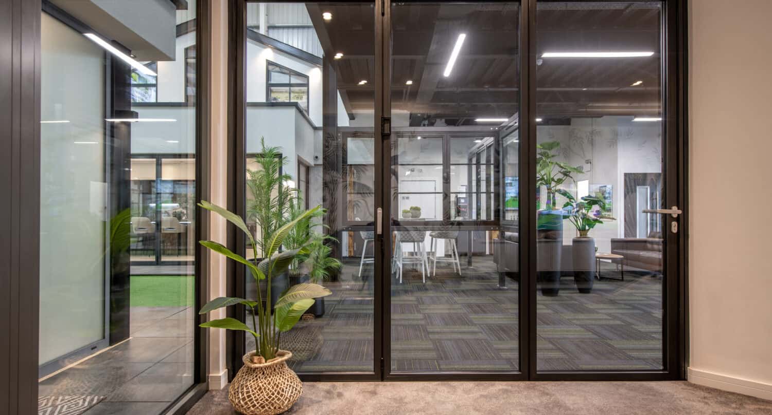 Modern office space with glass walls and bifold doors, indoor plants, and a view into a meeting area with tables, chairs, and contemporary decor.