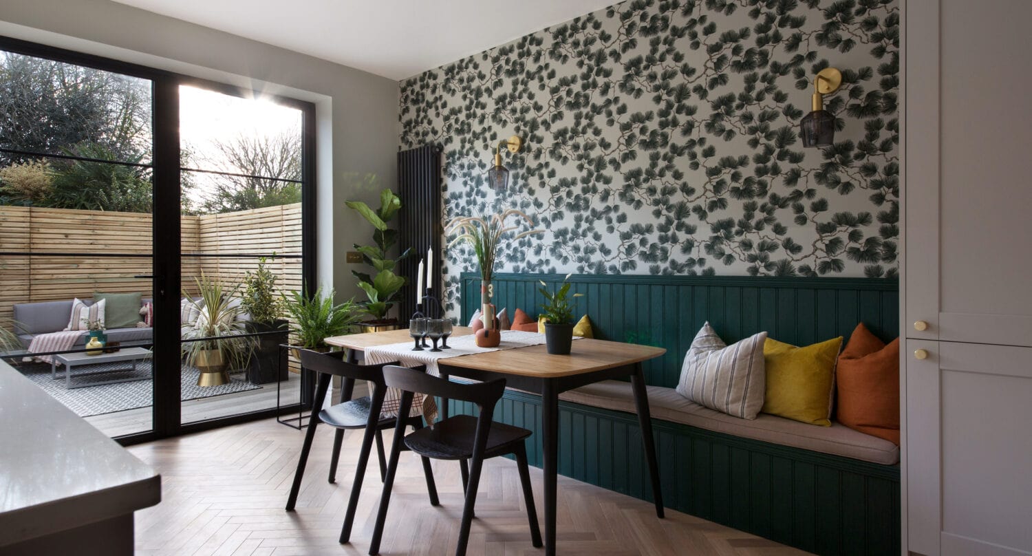 Modern dining area with a wooden table, black chairs, and a built-in bench with colorful cushions. Green leafy wallpaper and indoor plants decorate the space. Large bifold doors open to a sunny patio with outdoor seating.