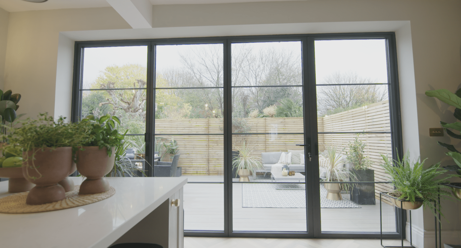 Large bifold doors open to a modern outdoor deck with seating, potted plants, and a wooden fence. Indoor plants sit on a white kitchen counter in the foreground, while trees provide a scenic backdrop.