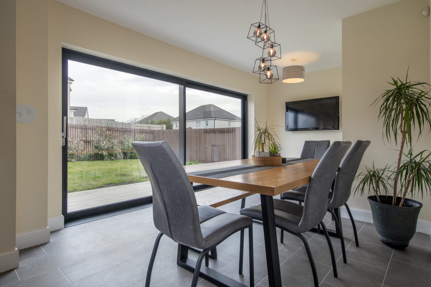 Modern dining room with a wooden table, six gray chairs, large sliding glass doors featuring bespoke glass solutions overlooking a backyard, potted plants, a wall-mounted TV, and a geometric ceiling light fixture.
