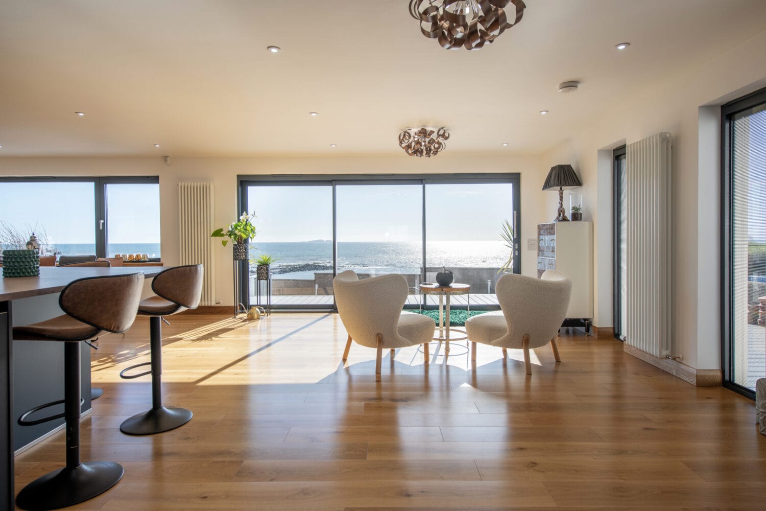 A bright, modern living space with large windows and bifold doors overlooking the ocean features wooden floors, two cozy chairs around a small table, and bar stools at a counter. Sunlight floods the room, creating a warm and inviting atmosphere.